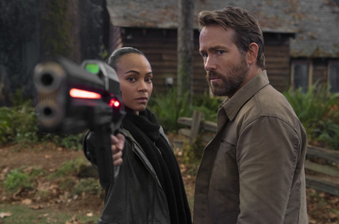 THE ADAM PROJECT Zoe Saldana as Laura holding laser rifle and Ryan Reynolds as Big Adam