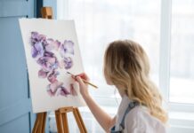 Creativity Boost: Tips To Help Improve Your Painting A woman in denim overalls and a white shirt is painting purple flowers onto a canvas that sits on a wooden easel.