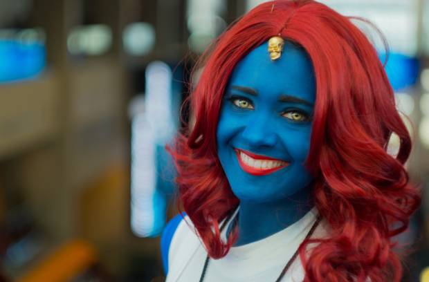 A young woman has blue skin, red hair, and red lipstick. She's wearing a white shirt, and a skull dangles on her forehead. A young woman has blue skin, red hair, and red lipstick. She's wearing a white shirt, and a skull dangles on her forehead.