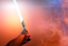 6 Geeky Gifts That Are Perfect To Give Star Wars Fans A person holding an illuminated orange light saber with two hands at the hilt. The background is foggy.
