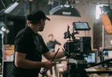 4 Single-Camera Sitcoms That Changed the TV Game A camera operator sets up a video camera on a set. In the background, an actor stands on set beneath the lights.