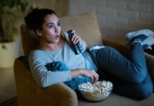 5 Best Ways To Stream Movies in Your Home A surprised woman sitting in an orange chair with a TV remote in her hand and a bowl of popcorn in front of her.