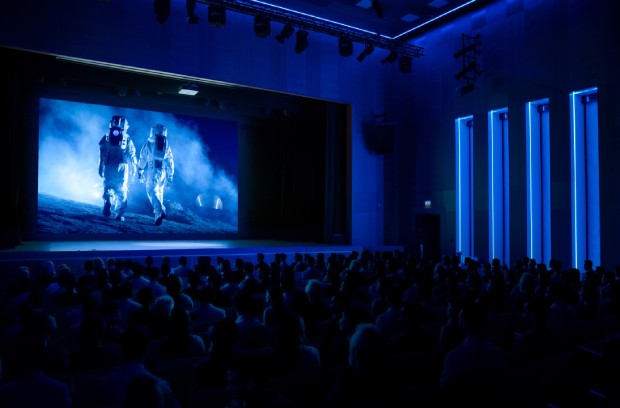 A dark theater with blue ambient lighting highlighting a large audience. The screen depicts two astronauts.