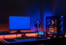 4 Strategies for Keeping Your Gaming Room Cool A gaming setup featuring a monitor displaying a blue screen and a colorful keyboard sit on a desk in a darkened room.