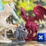 Materials You Can Try For DIY DnD Miniatures Three 3D printed miniatures of two dragons and a mage placed beside two dice on a map, all set against a detailed background.