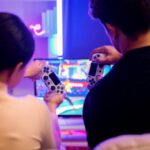 How To Build a Functional Gaming Space Anywhere A guy and girl sitting close to each other on the couch while holding gaming controllers. They are playing a fighting game.