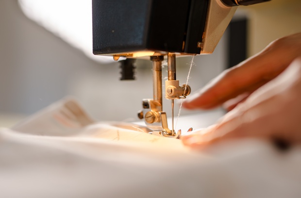 A close-up view shows a person pushing white fabric through the sewing needle of a sewing machine. A close-up view shows a person pushing white fabric through the sewing needle of a sewing machine.