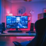 How To Build a Gaming Room on a Tight Budget A modern desk with a gaming chair in a colorfully lit room. The desk has a PC desktop, speakers, and gaming headphones.