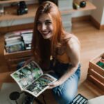 Essential Tips for Moving Your Comics and Collectibles A smiling woman with reddish hair sits on a wooden floor as she holds an open comic book. Crates of comics sit behind her.