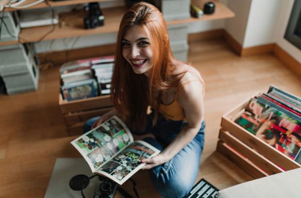 capitolnorthamerican-402821-woman-reading-comics-image1 A smiling woman with reddish hair sits on a wooden floor as she holds an open comic book. Crates of comics sit behind her.