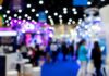 First-Time Comic Con Vendor Survival Tips A blurred view of a busy convention hall with colorful booths, bright lights, and crowds walking along a blue carpet.