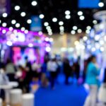 First-Time Comic Con Vendor Survival Tips A blurred view of a busy convention hall with colorful booths, bright lights, and crowds walking along a blue carpet.