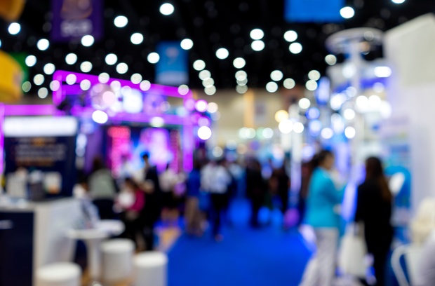 First-Time Comic Con Vendor Survival Tips A blurred view of a busy convention hall with colorful booths, bright lights, and crowds walking along a blue carpet.